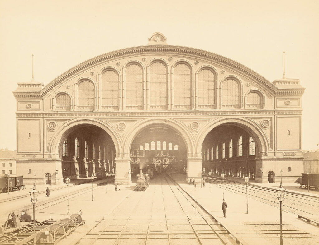Blick in eine monumentale Bahnsteighalle mit drei großen Rundbögen und einer abfahrenden Dampflok.