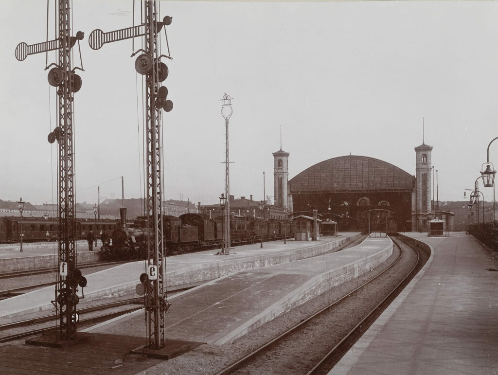 Blick von den Gleisen auf die Haupthalle, einem zentralen, dreigeteilten Hauptbau mit einem mittleren, erhöhten Gebäudeteil und zwei seitlichen Flügelbauten.