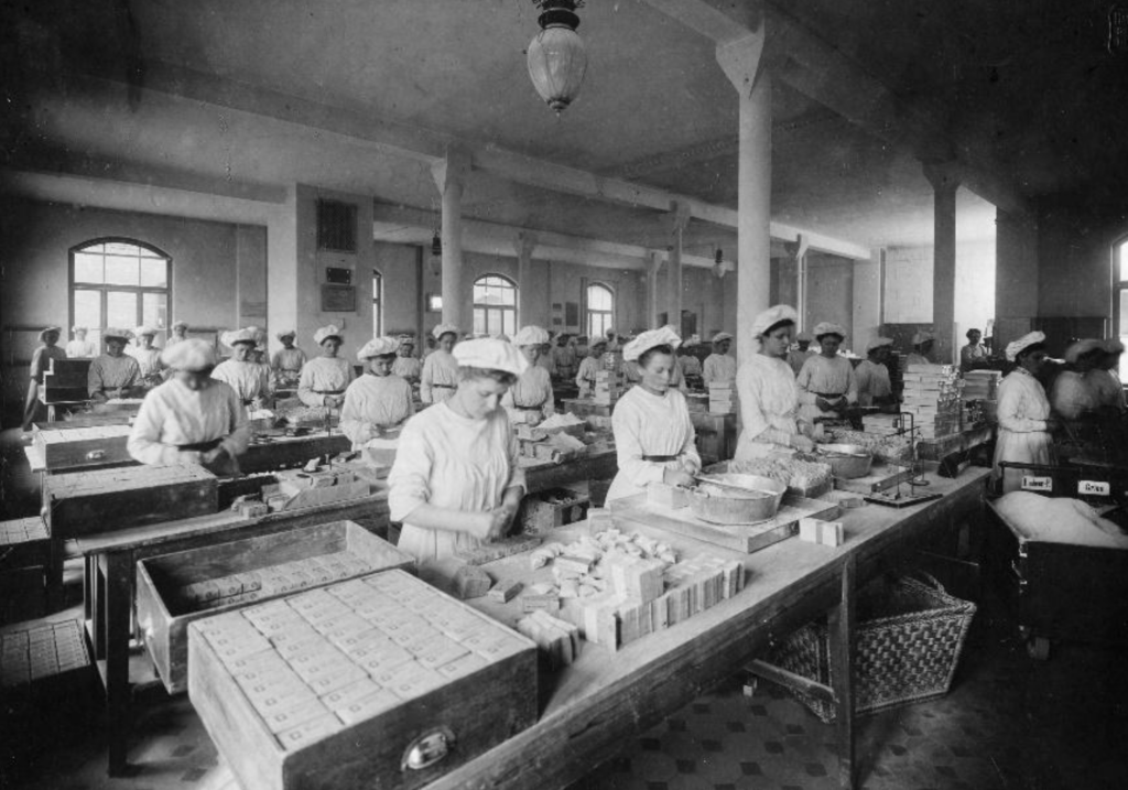 Frauen mit Hauben und Kitteln stehen in einer Fertigungshalle mit langen Tischreihen und füllen das Produkt ab.
