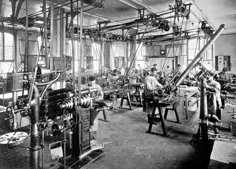 Produktionshalle, in der Männer in Kitteln an diversen Maschinen arbeiten.