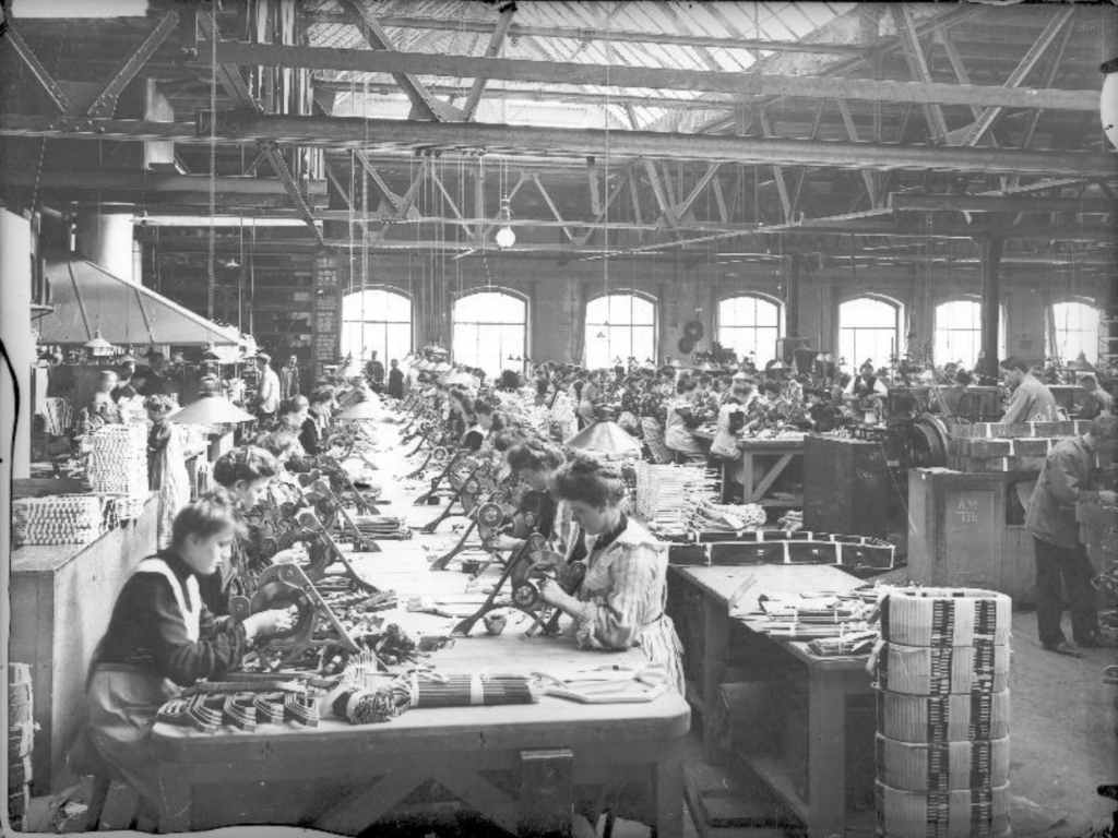 Arbeiterinnen sitzen in einer monumentalen Fertigungshalle an meterlangen und breiten Tischen und wickeln Spulen.