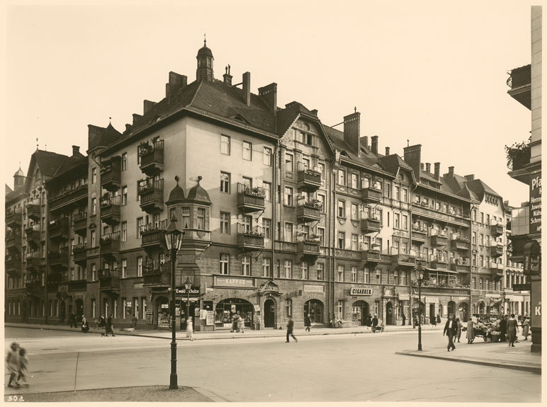 Straßenecke mit einem Wohnhaus. Im Erdgeschoss Rundbögen mit Schaufenstern und Werbetafeln von Läden.