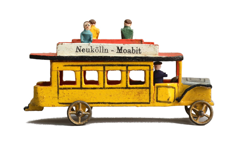 Gelber Spiel-Bus mit offener Sitzfläche auf dem Dach, auf der drei Figuren angedeutet sind. Aufschrift auf dem Dach: "Neukölln-Moabit".