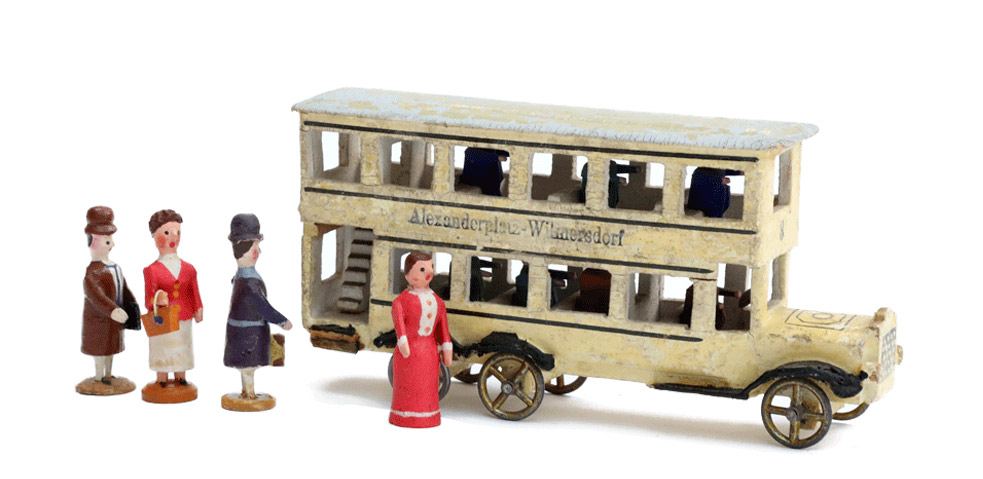 Miniatur eines Busses mit der Aufschrift "Alexanderplatz-Wilmersdorf." Es handelt sich um einen gelblichen Doppeldecker. Davor vier Spielfiguren in Form von Frauen.