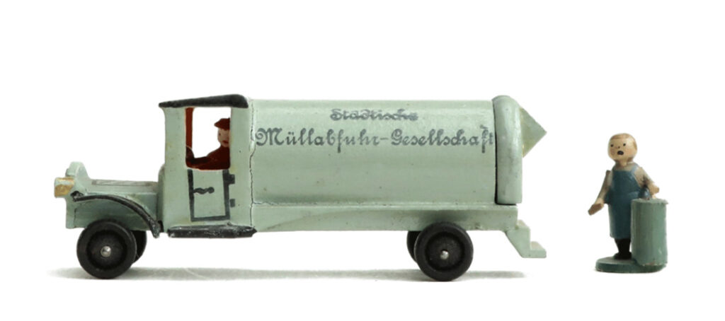 Zylinderförmiges Fahrzeug-Modell mit Fahrerfigur und Aufschrift: "Städtische Müllabfuhr-Gesellschaft." Der Wagen ist blassgrün, die Figur in rot-braun gekleidet. Neben dem Fahrzeug steht die Figur eines Arbeiters der Müllabfuhr mit ausdrucksstarkem, aufgemaltem Gesicht, der eine blassgrüne Mülltonne hält.