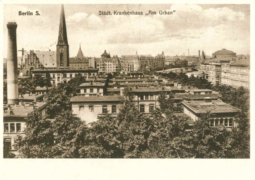 Leichte Vogelperspektive auf Dächer, Schornsteine und einen Kirchturm zwischen üppiger Begrünung. Aufschrift: "Berlin S. - Städt. Krankenhaus Am Urban."
