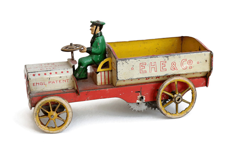 Farbenfrohes Blechmodell eines Lastwagens mit einem Männchen auf dem Fahrersitz. Unter dem mit der Wagen sind Zahnräder zu sehen. Aufschrift auf der Ladefläche: „EHE & Co.“. Aufschrift auf der Motorhaube: „ENGL. PATENT“.