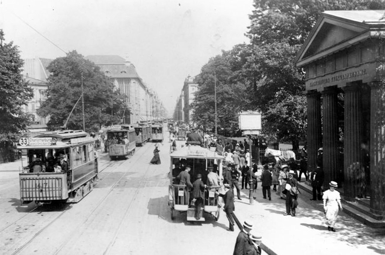 Ein klassizistisches Säulenportal neben der mit historischen Straßenbahnen befahrenen Straße.Im Hintergrund stehen zahlreiche Hochhäuser. Auf dem Bürgersteig laufen Passanten mit Strohhüten.