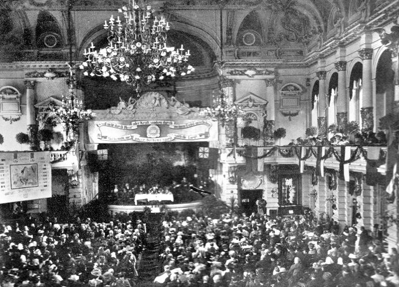 Blick in einen Theatersaal mit einer kleinen Bühne und eindrucksvollem Kronleuchter.